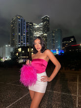 Woman styling a hot pink ostrich feather evening bag with a pink satin top and white mini skirt against a nighttime city skyline